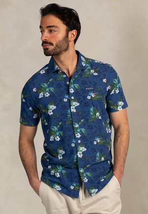 Man met baard die een blauw kort mouw overhemd draagt met een groen-wit bloemenpatroon en beige broek, handen in de zakken, kijkt naar links.