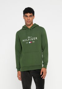 Hoodie vert avec une poche avant, présentant le logo "Tommy HILFIGER" imprimé en blanc. Tissu doux et texturé avec une capuche à cordon.