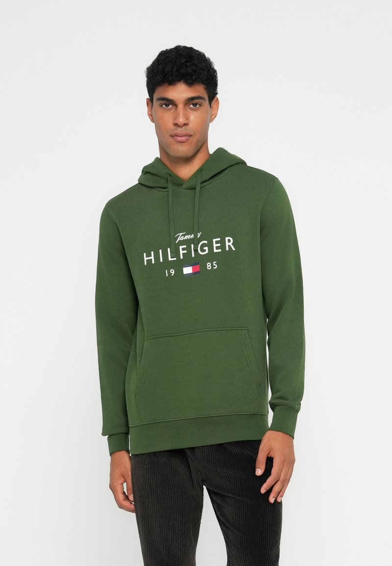 Hoodie vert avec une poche avant, présentant le logo "Tommy HILFIGER" imprimé en blanc. Tissu doux et texturé avec une capuche �à cordon.