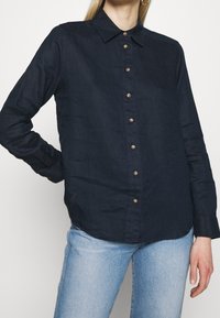 Camicetta blu navy con bottoni, realizzata in tessuto leggero, dotata di colletto e bottoni in tonalità oro, con maniche lunghe e orlo leggermente arrotondato.