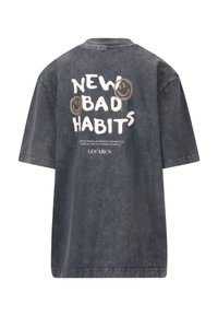 Yli kokoa tumma farkkupaita, jossa on rohkea valkoinen ja kullanvärinen teksti "UUSI HUONO TAPA" sekä hymyileviä kasvoja graafisena kuviona selkämyksessä. Lyhyet hihat, rento istuvuus.