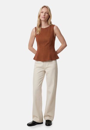 Vrouw staand met handen achter de rug, draagt een mouwloze bruine top, crème wijde broekspijpen en zwarte loafers op een witte achtergrond.