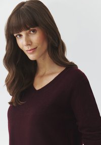 Maglione bordeaux a scollo a V con una consistenza morbida, dotato di maniche lunghe e vestibilità rilassata, indossato da un modello con capelli lunghi fino alle spalle.