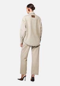 Naine seljaga seistes, seljas beež ülev suuruse teksajakk ja sobivad püksid, tumedast pruunist kõrge kontsaga kingad, juuksed kogutud bun'i.