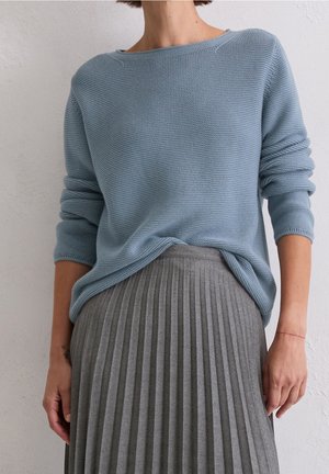 Femme portant un pull en maille bleu clair avec les manches retroussées et une jupe plissée grise taille haute, debout contre un mur blanc.