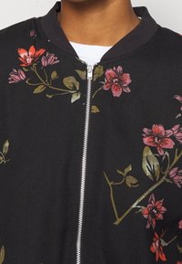 Veste bomber noire avec un motif floral aux accents roses, rouges et dorés, fermeture éclair et tissu texturé.