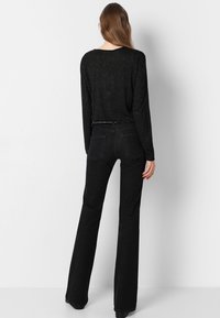 Haut noir à manches longues avec des accents pailletés, associé à un jean noir évasé. La tenue présente une taille ajustée et deux poches arrière sur le jean.