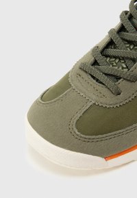 Olivově zelená teniska kombinující semišový a síťovinový materiál. Gumová podrážka s oranžovým akcentem, kulatá špička a texturovaná oblast šněrování.