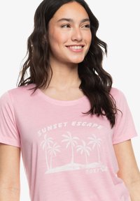 Lichtroze T-shirt met korte mouwen, voorzien van een witte palmboomeGraphic en de tekst "SUNSET ESCAPE" in een gebogen ontwerp. Zachte textuur.
