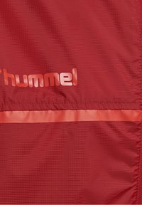 Tissu rouge texturé avec une légère bande horizontale en relief et le logo rouge brillant "hummel" partiellement visible.
