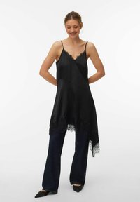 Tunique en satin noir avec un ourlet en dentelle, dotée d'un col en V et de fines bretelles. Associée à un jean en denim foncé et des chaussures noires à bout pointu.