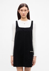 Μαύρο φόρεμα καπιτονέ με τετράγωνη λαιμόκοψη, δύο φαρδιές τιράντες και μπροστινή τσέπη με λευκό κείμενο. Λευκό πουκάμισο με μακριά μανίκια από κάτω.