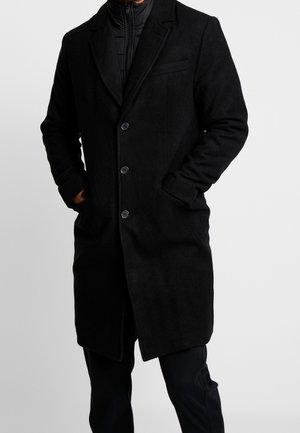 Homme portant un manteau en laine noir mi-long à trois boutons, les mains dans les poches, par-dessus une veste matelassée noire zippée et un pantalon noir.