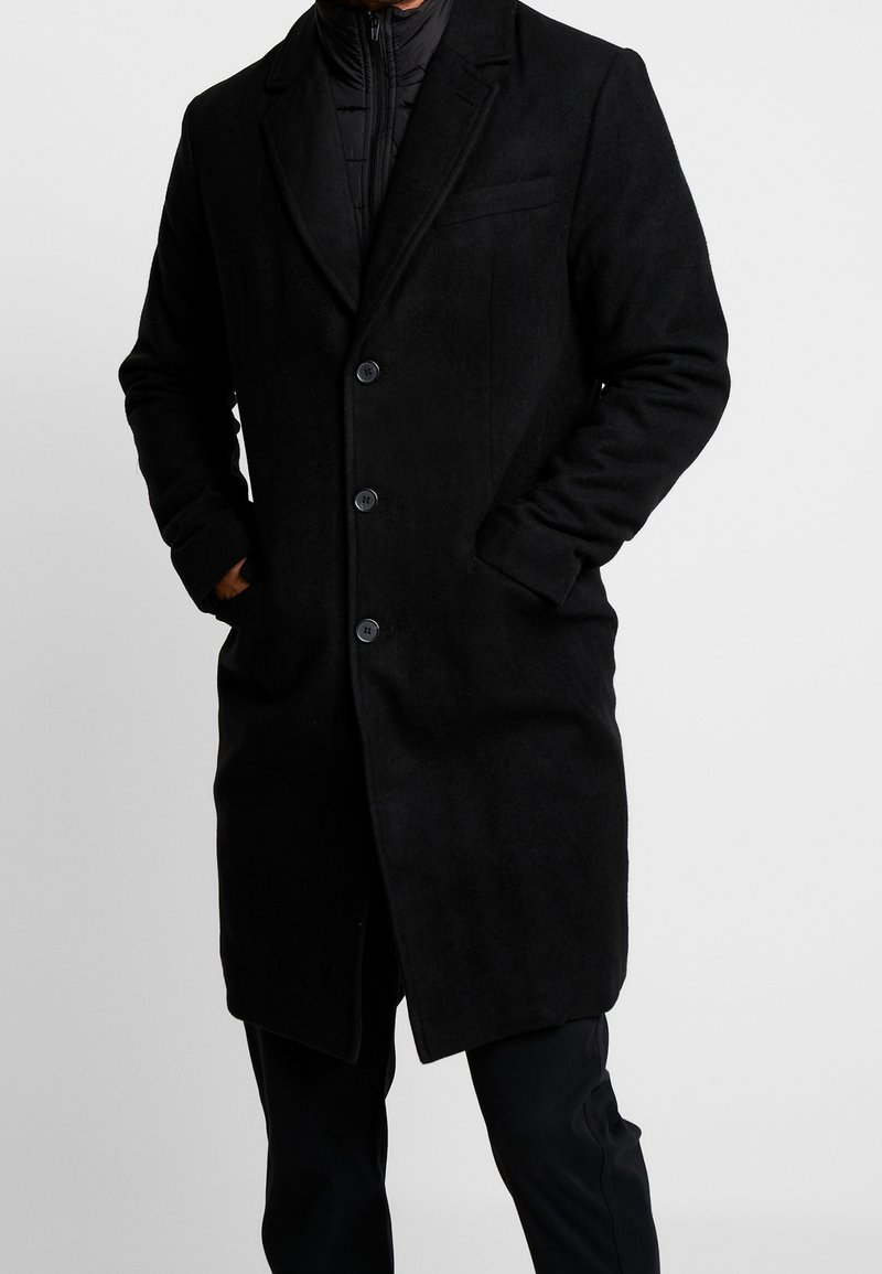 Homme portant un manteau en laine noir mi-long à trois boutons, les mains dans les poches, par-dessus une veste matelassée noire zippée et un pantalon noir.