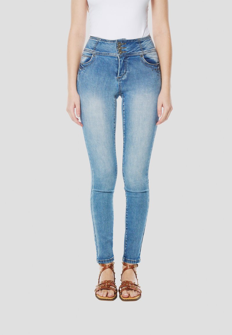 Blauwe denim skinny jeans met een hoge taille, voorzakken en decoratieve knoopdetails. Gecombineerd met bruine sandalen met studs.