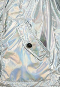 Tissu de veste argenté irisé avec une poche diagonale ornée d'un bouton noir et d'une couture contrastante. Texture lisse et brillante.