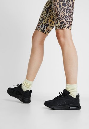 Jambes portant des chaussettes jaunes, des chaussures de sport noires et un short à imprimé léopard sur un fond clair uni.
