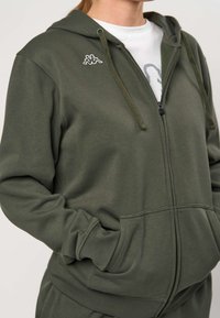 Person trägt olivgrünen Kapuzenpullover mit Reißverschluss, Fronttaschen und kleinem weißem Logo auf der Brust, Hände in den Taschen, über einem weißen Hemd.