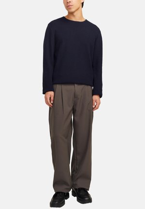 Uomo che indossa un maglione lungo a maniche nere scure, un paio di pantaloni marroni a pieghe larghi e scarpe in pelle nere, in piedi su uno sfondo semplice.