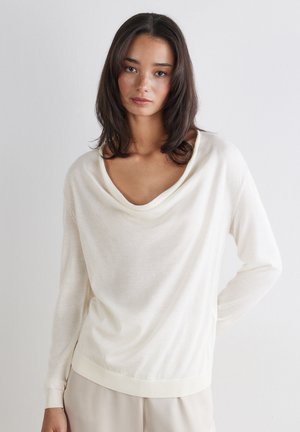 Junge Frau mit dunkelbraunem, schulterlangem Haar, die ein locker sitzendes weißes Langarmshirt trägt und vor einem schlichten hellen Hintergrund steht.