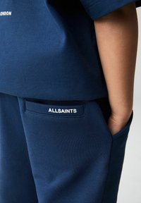 Mornarsko plava pamučna košulja kratkih rukava s okruglim izrezom i vidljivim logotipom "ALLSAINTS". Prateće mornarsko plave hlače s detaljem na džepu.