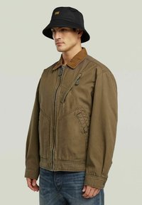 Veste zippée marron avec un col en velours côtelé, dotée de deux poches avant et d'un détail de fermeture éclair. Portée avec un chapeau seau noir.