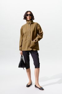 Naine, kellel on seljas pruun nööpidega jope, mustad lühikesed püksid, mustad madalad jalanõud, päikeseprillid, kandes musta käekotti, seisab valge tausta ees.