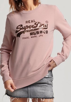 Kvinde iført en pink sweatshirt med teksten "SuperDry" i palietter og et falmet denimskørt med frynset kant, poserer med den ene hånd i lommen.