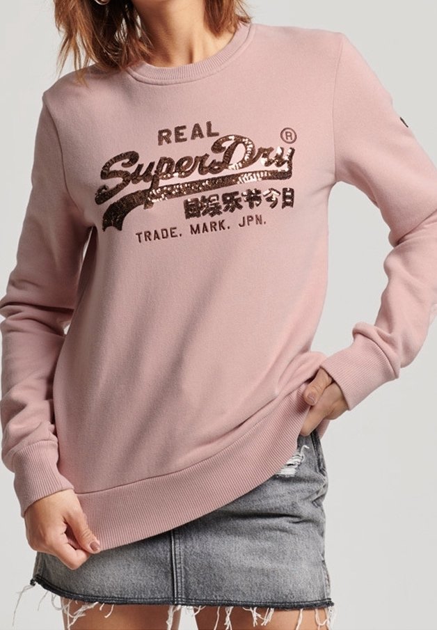 Femme portant un sweat rose avec le texte "SuperDry" en sequins et une jupe en jean délavée à ourlet effiloché, posant avec une main dans la poche.