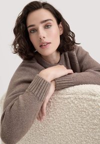 Ruskea neulottu villapaita, jossa on ribbireunat, lepäävän teksturoidulla vaalean kermaisen pinnan päällä. Pehmeä, mukava materiaali ja rento muotoilu.