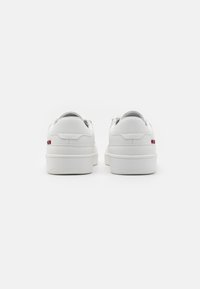 Adidași albi cu o textură de piele netedă, spate rotunjit, talpă plată din cauciuc și accente subtile de roșu și albastru lângă călcâi.