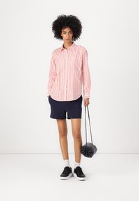 Red and white striped button-up shirt paired with navy shorts, black sneakers, and a small dark bag. The shirt has a chest pocket.
