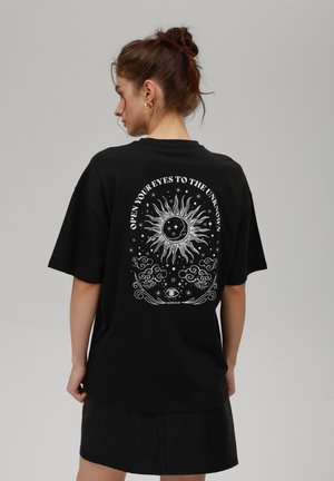 Black cotton t-shirt featuring a large white sun design with celestial elements and text "OPEN YOUR EYES TO THE UNKNOWN" on the back.