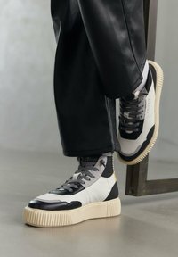Sneakers alte con pelle nera e bianca, dettagli grigi testurizzati e una suola spessa color crema. Design con lacci e dettaglio del logo visibile.
