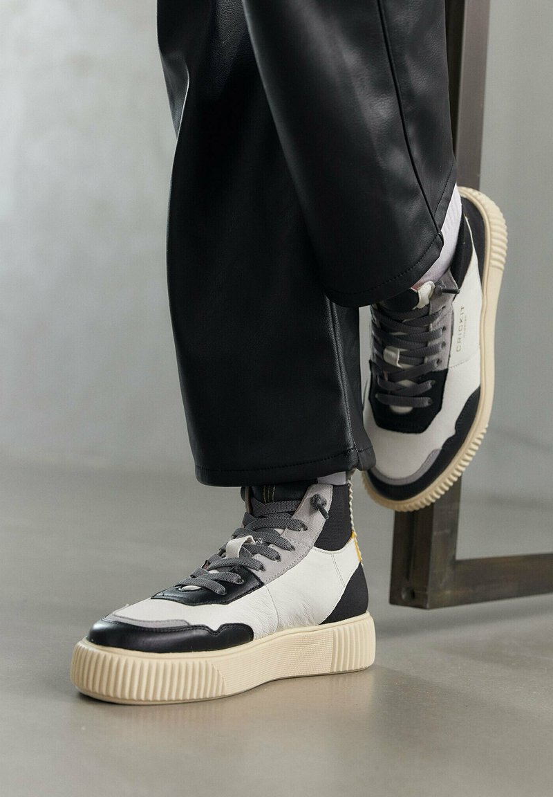 Sneakers alte con pelle nera e bianca, dettagli grigi testurizzati e una suola spessa color crema. Design con lacci e dettaglio del logo visibile.