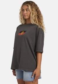 Grijze oversized T-shirt met korte mouwen, voorzien van een opvallend ontwerp van een zwart, rood en gouden cirkelpatroon aan de voorkant. Gecombineerd met denim short.