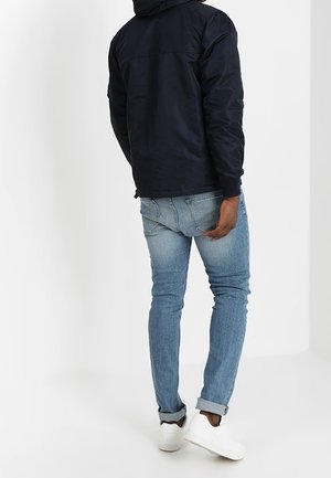 Mann trägt eine schwarze Jacke, hellblaue hochgekrempelte enge Jeans und weiße Sneaker, steht mit dem Rücken zur Kamera.