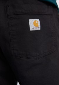 Juodos kelnės su galiniu kišeniu, kuriame yra baltas etiketė. Etiketėje pavaizduotas geltonas logotipas ir raudonas tekstas, nurodantis prekės ženklą "Carhartt."