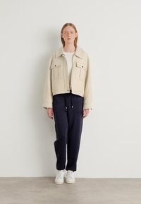 Giacca beige cropped con due tasche sul petto, indossata su una maglietta bianca, abbinata a joggers blu scuro e sneakers bianche. Texture liscia, vestibilità casual.