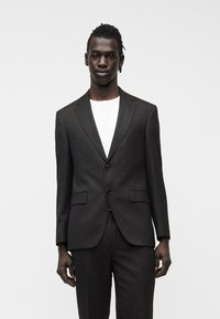 Νεαρός άνδρας με σκουρόχρωμο δέρμα και πλεξούδες, που φοράει σκούρο καφέ σακάκι και παντελόνι πάνω από λευκό μπλουζάκι, στέκεται μπροστά σε λευκό απλό φόντο.