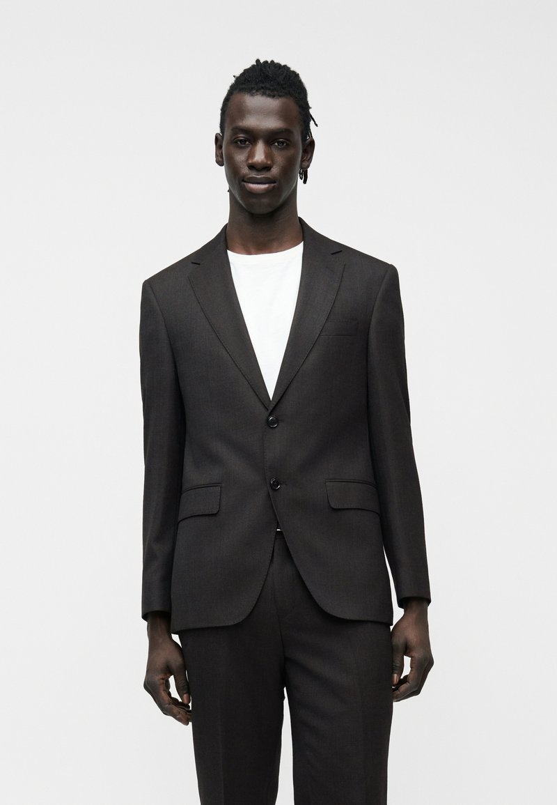 Νεαρός άνδρας με σκουρόχρωμο δέρμα και πλεξούδες, που φοράει σκούρο καφέ σακάκι και παντελόνι πάνω από λευκό μπλουζάκι, στέκεται μπροστά σε λευκό απλό φόντο.