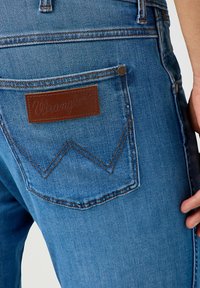 Jeans de mezclilla azul con una etiqueta de cuero marrón que presenta el logo "Wrangler". Detalle de costura notable en los bolsillos traseros.