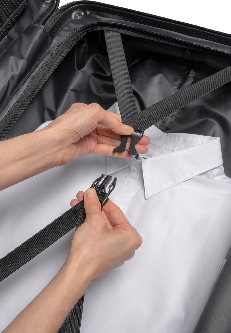 Zwarte stoffen kofferinterieur met een wit overhemd, vastgezet met zwarte webbingriemen en plastic clips. Handen passen de riemen aan.