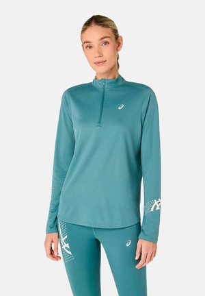 Vrouw draagt teal lange mouwen sporttop met kwartrits en bijpassende leggings met witte merklogo's op mouw en dij.