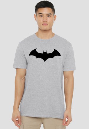 Man wearing light gray T-shirt with large black Batman logo centered on chest, standing against plain background.