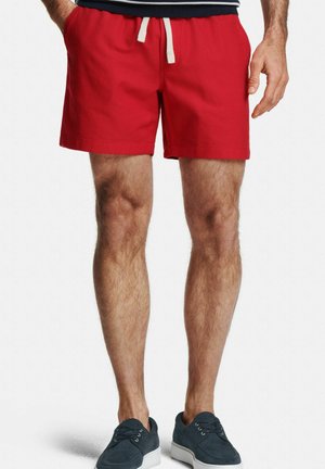Mann trägt rote Shorts mit Kordelzug und marineblaue legere Schnürschuhe, steht mit teilweise sichtbaren Händen.