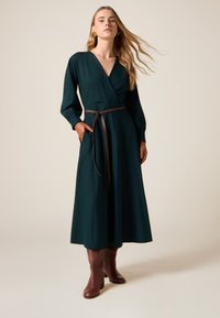 Dlouhé tmavě zelené zavinovací šaty z hladké látky, s dlouhými rukávy, hnědým páskem v pase a bočními kapsami. Nošeny s hnědými kotníkovými botami.