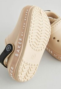 Zapatos tipo zueco beige con suela texturizada y correa ajustable negra. "BAKER by TED BAKER" está grabado en blanco en el lateral.