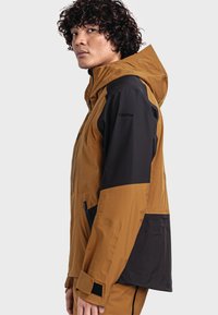 Braune und schwarze wetterfeste Jacke mit Kapuze, ausgestattet mit einem Farbschnittdesign, Reißverschlusstaschen und verstellbaren Bündchen für einen sicheren Sitz.