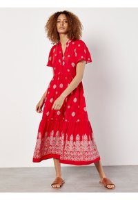 Rotes Maxikleid mit kurzen Ärmeln, V-Ausschnitt und Knopfdetails. Mit weißem Blumenmuster und gestuftem Rock; getragen mit braunen Rutschsandalen.